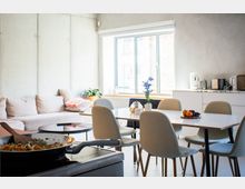 Heller, moderner Wohn- und Essbereich mit großem Fenster, hellem Sofa und einem rechteckigen Esstisch mit mehreren gepolsterten Stühlen. Rechts befindet sich eine schlichte weiße Küchenzeile mit kleinen Küchengeräten und Arbeitsfläche.