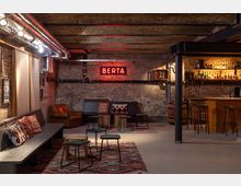Gemütlicher Barbereich in einem rustikalen Kellerraum mit unverputzten Ziegelwänden, freiliegenden Rohren und einer roten Neonleuchtschrift „BERTA“. Der Raum ist mit gemusterten Teppichen, Polstermöbeln und einer Bar aus Holz mit Flaschenregal ausgestattet.