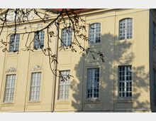 Gelbe Fassade eines historischen Gebäudes mit großen Sprossenfenstern und dekorativen Stuckelementen unterhalb des Dachs; im Vordergrund sind Zweige eines Baumes zu sehen, deren Schatten auf der Wand erkennbar sind.