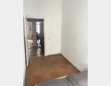 Ein kleiner Raum mit weiß gestrichenen Wänden und braunem Bodenbelag aus Kork. Im Hintergrund führt eine offene Tür in einen anderen Raum mit Holzboden, einem Fahrrad und einer Topfpflanze.