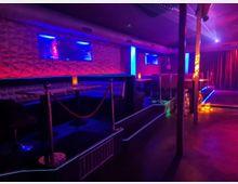 Innenraum eines Nachtclubs mit dunkel gepolsterten Sitznischen und Tischen entlang einer strukturierten Wand, beleuchtet von blauen und violetten Neonstreifen. Rechts befindet sich ein kleiner Bühnenbereich mit vertikalen Stangen und roten Vorhängen, ergänzt durch Akzentlichter und Seilabsperrungen.