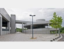 Betonierter Parkplatz mit modernem Gebäudekomplex, bestehend aus geschwungenen, offenen Betonrampen und einem Aufbau mit hohen Pfeilern, daneben ein einzelner Baum. Der Bereich ist teils von Straßenlaternen und gepflastertem Boden umgeben, im Hintergrund sind Pflanzen und Bäume sichtbar.