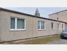 Flacher Plattenbau mit einer rauen, beigen Fassade und mehreren rechteckigen Fenstern mit weißen Rahmen und halbtransparenten Gardinen. Im Vordergrund steht ein graues Auto, im Hintergrund sind ein Baum und ein weiteres Gebäude sichtbar.