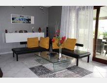 Modernes Wohnzimmer mit einer schwarzen Leder-Sitzbank, dekoriert mit gelben Kissen. Im Vordergrund steht ein rechteckiger Glastisch mit einer roten Blumenvase, während im Hintergrund eine weiße Wandkommode und ein großes Fenster mit transparenten Vorhängen zu sehen sind, das auf einen Balkon führt.