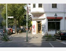 Eine Straßenecke in einer Stadt mit einem Gebäude, auf dem „Antiquariat“ steht. Vor dem Geschäft stehen Fahrräder und ein Verkehrsschild, daneben sind eine Fußgängerampel, geparkte Autos sowie Bäume im Hintergrund zu sehen.