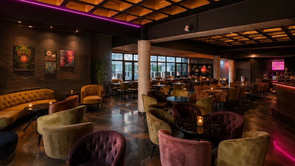 Innenraum einer modernen Lounge-Bar mit vielen runden Tischen und gepolsterten Samtsesseln in verschiedenen Farben, auf einem glänzenden Boden. Dunkle Wände mit Kunstbildern, große Fensterfronten, Säulen sowie eine kassettierte Decke mit warmem Licht und violetten LED-Akzenten prägen den Raum.