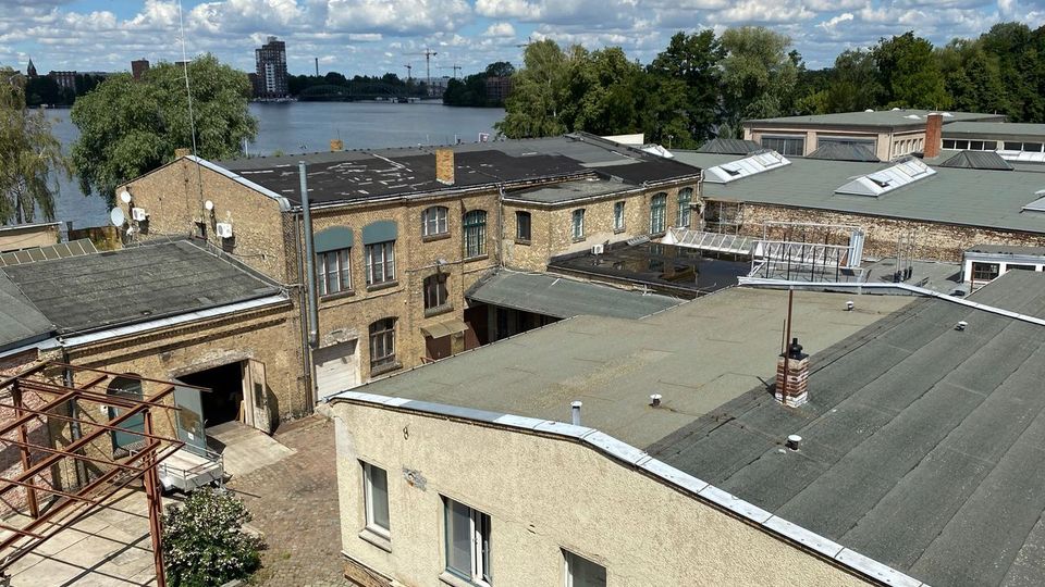 Blick über flache Dächer und Backsteingebäude eines ehemaligen Industrie- oder Lagerkomplexes mit Innenhof und Oberlichtern. Im Hintergrund liegt ein breiter Fluss oder See mit Baumufer und einer entfernten Stadtsilhouette unter wolkigem Himmel.