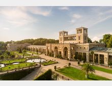 Historisches Gebäude mit klassizistischer Architektur, darunter Säulen, Bögen und Statuen, umgeben von weitläufigen Gartenanlagen mit dekorativen Teichen, Rasenflächen und Pflanzen. Im Hintergrund ist ein bewaldeter Bereich und ein blauer Himmel sichtbar.