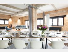 Loftartiger Raum mit unverputzten Backsteinwänden, großen Fenstern und Sichtbetondecken. Im Vordergrund steht ein gedeckter Esstisch mit weißen Stühlen, im Hintergrund eine Sitzecke mit Sofas und moderner Dekoration.