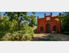 Backsteingebäude im neugotischen Stil mit drei Spitzbogenfenstern und einem Uhrturm, umgeben von dichtem Grün und Bäumen; ein kleiner Pfad mit Stufen führt zu dem Eingang.