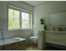 Modernes Badezimmer mit einer Badewanne mit Mosaikverkleidung, einem weißen Waschbecken mit Spiegel darüber und einem Fenster mit Blick ins Grüne. Die Wände sind mit weißen Fliesen verkleidet, der Boden ist mit hellen Fliesen gestaltet.