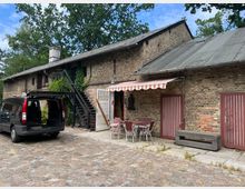 Backsteinbau mit einem Ziegeldach, einer Außentreppe und rot-weiß gestreifter Markise über einem Sitzbereich mit Eisenmöbeln. Im Vordergrund ein geöffneter schwarzer Van auf einem gepflasterten Hof, umgeben von Bäumen.