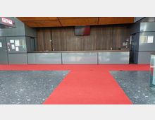 Eine Empfangshalle mit roten Teppichen, einem dunklen Holzhintergrund, einer langen Empfangstheke und grauen Wandverkleidungen. Links sind Schilder wie „Sprinklerzentrale“ sowie Notfallhinweise sichtbar.