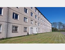 Plattenbau mit vier Etagen und zahlreichen rechteckigen Fenstern, einige davon vergittert; umgeben von einer Rasenfläche und Bäumen im Hintergrund, bei klarem, blauem Himmel.