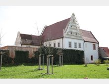 Historisches Gebäude mit steilem Giebeldach und kunstvollen Stufengiebeln in einer umliegenden Grünfläche mit jungen Bäumen. Eine mit Efeu bewachsene Mauer und ein Holzzaun grenzen den Gartenbereich ab.