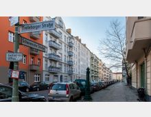 Straßenszene mit mehrstöckigen Wohngebäuden im Altbaustil, teils mit Balkonen in Pastellfarben. Links Straßenschilder für die "Trelleborger Straße" und "Schonensche Straße", geparkte Autos und ein Bürgersteig mit einem Baum und einer Wasserpumpe.