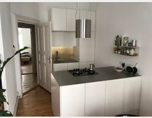 Kleine, moderne Küche mit weißen Einbauschränken, einer Edelstahl-Arbeitsplatte und einer Dunstabzugshaube. Im Hintergrund führt eine offene Tür in ein angrenzendes Zimmer mit Holzfußboden.