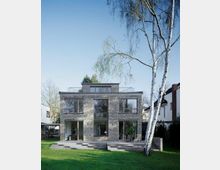 Moderne zweistöckige Wohnhausfassade aus grauem Ziegelstein mit großen Glasfenstern, einer Terrasse und einer Treppe, umgeben von einem grünen Rasen und hohen Bäumen, darunter eine schlanke Birke im Vordergrund.