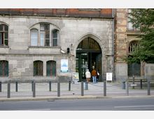 Außenansicht eines historischen Gebäudes mit Stein- und Klinkerfassade sowie großen, gotischen Rundbogenfenstern. Der Haupteingang mit einem großen, spitzbogigen Portal ist verglast, davor stehen zwei Personen; Schilder und Plakate weisen auf eine Galerie hin.