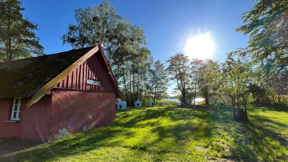 Kleines rotes Holzhaus mit steilem, moosbewachsenem Satteldach am Rand einer grünen Wiese, umgeben von hohen Bäumen. Im Hintergrund sind mehrere kleine weiße Häuschen aufgestellt, dahinter glitzert ein See unter tief stehender Sonne am klaren Himmel.