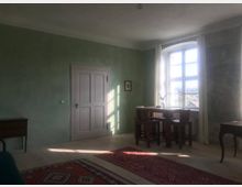 Ein schlicht eingerichtetes Zimmer mit grün gestrichenen Wänden, einem Tisch und vier Holzstühlen vor einem großen Fenster mit weißen Vorhängen. Der Boden ist mit hellen Holzdielen und roten Teppichen bedeckt, und links steht eine Tür.