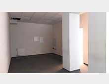 Ein leerer Raum mit hellen Wänden, grauem Teppichboden und einer Decke mit Rasterplatten. An der linken Wand befindet sich ein Heizkörper, während an der Rückwand ein Wasseranschluss und kleine geflieste Flächen sichtbar sind.