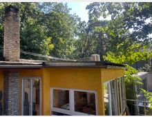 Gelbes Gebäude mit Putz in Schuppenmuster-Optik, großen Fenstern und einem Schornstein aus Backstein. Das Gebäude ist von dichtem Grün umgeben, einschließlich Bäumen und Sträuchern, und ein weiterer Gebäudeteil ist im Hintergrund erkennbar.