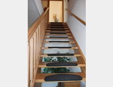 Holztreppe mit schwarzem Teppich auf den Stufen, flankiert von einem Holzgeländer auf der linken und einem Wandhandlauf auf der rechten Seite. Am oberen Ende der Treppe befindet sich eine offene Tür, durch die ein heller Raum mit einer Blumenvase auf einem Tisch sichtbar ist.