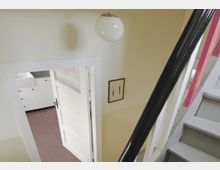 Blick in einen Innenraum mit einer offenen Tür, die zu einer weißen Küche mit weißen Schränken und einer Spüle führt. Eine Treppe mit schwarzem Geländer und grauen Stufen sowie eine beige Wand mit einem Lichtschalter und einer Deckenleuchte sind zu sehen.