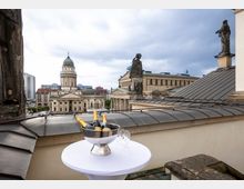 Blick von einer Dachterrasse auf den Gendarmenmarkt in Berlin mit dem Französischen Dom im Vordergrund und dem Konzerthaus im Hintergrund. Auf einem Stehtisch im Vordergrund stehen eine Metallschale mit Sektflaschen und mehrere leere Sektgläser.