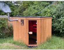 Eine kleine Sauna aus Holz mit einem abgewinkelten Schornstein, aus dem Rauch aufsteigt, steht in einer grünen Umgebung mit Bäumen und Sträuchern. Durch die Glastür ist ein Inneres mit roten Lichtern sichtbar.