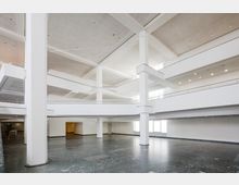 Großer, moderner Innenraum mit hohen Decken, weißen Wänden und Säulen sowie mehreren offenen Ebenen und Galerien. Der Boden besteht aus dunkelgrauem Stein, und große Fenster lassen Tageslicht in den Raum.