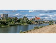 Flusslandschaft mit einer Uferpromenade, im Hintergrund eine rote Backsteinkirche mit spitzem Turm sowie eine moderne Brücke. Auf der linken Seite stehen ein Mehrfamilienhaus und kleinere Wohnhäuser, umgeben von Bäumen und Wiesen.