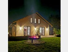 Ein Einfamilienhaus mit heller Fassade und geneigtem Dach, beleuchtet durch Wandlampen, bei Nacht. Im Vordergrund steht eine feuerschale auf einer Rasenfläche, umgeben von Pflanzen und einer Terrasse.