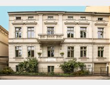 Vorderansicht eines historischen Gebäudes mit heller Fassade, dekorativen Architekturdetails und großen Fenstern. Vor dem Gebäude befindet sich ein kleiner Vorgarten mit Pflanzen, umgeben von einem schmiedeeisernen Zaun.