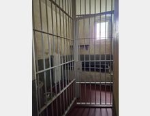 Zelle in einem Gefängnis mit vergitterter Tür, grau-beige Wänden, einer einfachen Liege, einem kleinen Fenster mit Gitterstäben und einer Metallschüssel auf dem Boden.