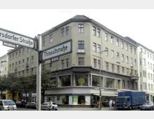 Ecke eines mehrstöckigen, beige verputzten Wohn- und Geschäftshauses mit großen Fenstern im Erdgeschoss, in denen sich Geschäfte wie ein Wellness-Studio befinden. Straßenschilder der Thrasoltstraße und Garsdorfer Straße sowie parkende Autos und ein blauer Lieferwagen sind im Vordergrund sichtbar.