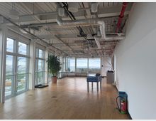 Spacious modern office lounge with floor-to-ceiling windows overlooking a city view, wood plank flooring, and an exposed industrial ceiling with ducts and piping. The open area includes a large potted plant, a gray sectional sofa, and a foosball table near a white wall.