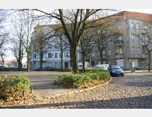 Gepflasterter Platz mit einzelnen Bäumen und niedrigem Buschwerk, umgeben von mehrstöckigen Wohnhäusern mit Putzfassaden und Balkonen. Am Straßenrand sind geparkte Autos und ein Wohnmobil zu sehen.