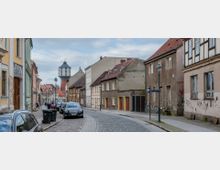 Straßenszene in einer Altstadt mit gepflastertem Straßenbelag, Reihenhäusern mit teils verputzten und teils Fachwerkfassaden sowie einem markanten Wasserturm im Hintergrund; parkende Autos säumen die Straße.