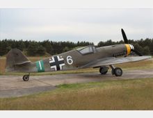 Ein historisches einmotoriges Jagdflugzeug aus dem Zweiten Weltkrieg, geparkt auf einem Betonvorfeld, mit einer grünen und olivfarbenen Tarnbemalung, einem Balkenkreuz-Symbol an der Seite sowie einer gelben Markierung auf dem Propellerbereich; im Hintergrund ist eine Wiese und Wald zu sehen.