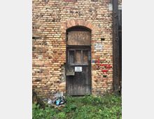 Eine alte Ziegelmauer mit einer verwitterten Holztür, die teilweise mit Brettern vernagelt ist. Auf der Tür hängt ein Schild mit der Aufschrift „Betreten verboten! Lebensgefahr!“, und im Vordergrund liegt Müll auf überwuchertem Gras.