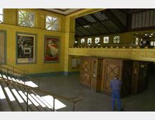 Ein historischer U-Bahnhof mit gelb gestrichenen Wänden, großen Fenstern im oberen Bereich und dekorativen Holzdetails. An den Wänden hängen Retro-Plakate, und Treppen führen zu verschiedenen Ebenen der Station.