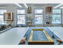 Ein Atelier mit großen Fenstern, das als Werkstatt für Bilderrahmen dient. Im Vordergrund liegt ein vergoldeter Rahmen auf einem Arbeitstisch, umgeben von Werkzeugen, während an den Wänden Regale mit Materialien und Bilderrahmen hängen.