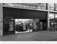 Schwarz-weiße Außenansicht eines modernen Ladengeschäfts mit großen Glasfronten und der Aufschrift „IMAGO“ auf mehreren Fenstern. Dahinter sind ein großer, zylindrischer Fotoapparat und verschiedene ausgestellte Objekte sichtbar.