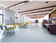 Großer und heller Speisesaal mit hohen Decken und sichtbaren Holzbalken. Der Raum ist mit bunten Stühlen und weißen Tischen möbliert, großen Fenstern an den Seitenwänden und einer Buffetstation aus dunklem Holz rechts im Vordergrund.