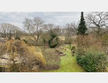 Weitläufiger Garten mit Rasenflächen, einer Feuerstelle in der Mitte, verschiedenen Bäumen und Sträuchern. Im Hintergrund sind karge Bäume und eine offene Landschaft unter bewölktem Himmel sichtbar.