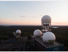 Ehemalige Abhörstation mit markanten weißen Kuppeln in Kugelform, die auf einem verlassenen Gebäude stehen; im Hintergrund ein dichter Wald und ein See bei Sonnenuntergang.