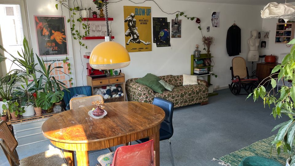 Eclectic living room with a round wooden dining table and mixed chairs in the foreground, a yellow dome pendant lamp, and a patterned sofa against a white wall covered with posters. The space features many potted and hanging plants, open shelving, and a carpeted floor with assorted vintage furniture and decor.