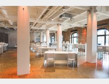 Großer, heller Veranstaltungsraum mit industriellem Charme, gekennzeichnet durch sichtbare weiße Deckenbalken, Backsteinwände und große Rundbogenfenster. Der Raum ist mit weißen Tischen und Stühlen sowie hohen Stehtischen ausgestattet, die auf einem Holzboden arrangiert sind.